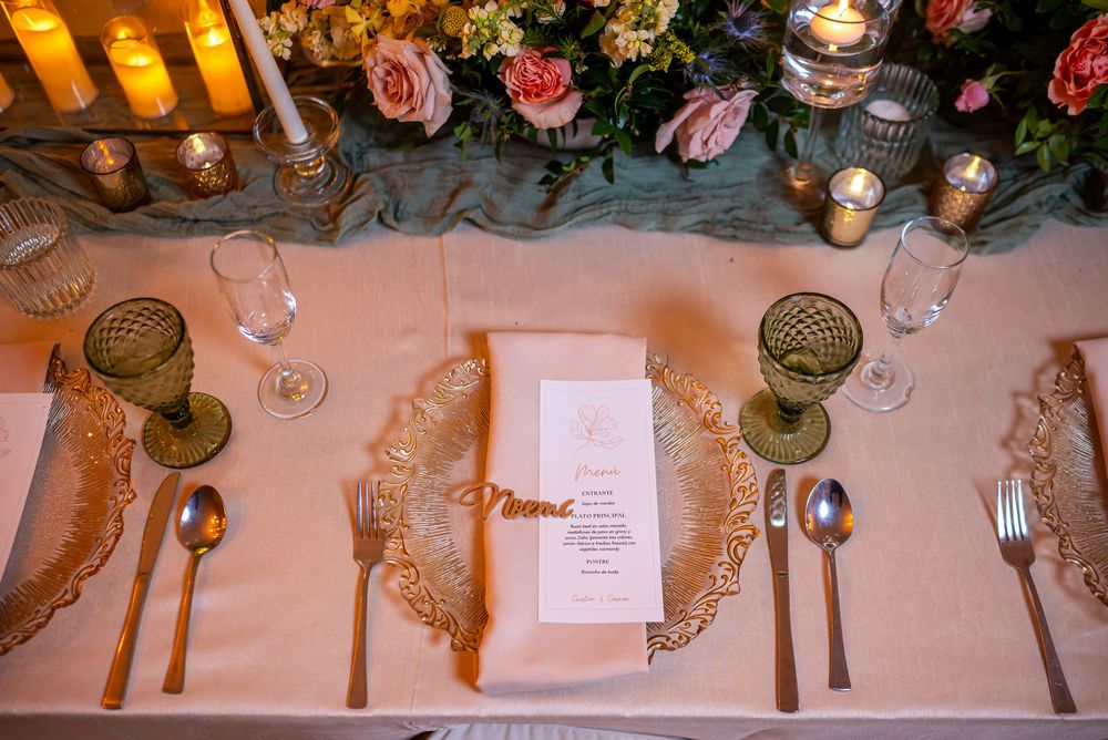 Elegant wedding place setting with gold charger plates, candles, pink roses and fine cutlery on white tablecloth.