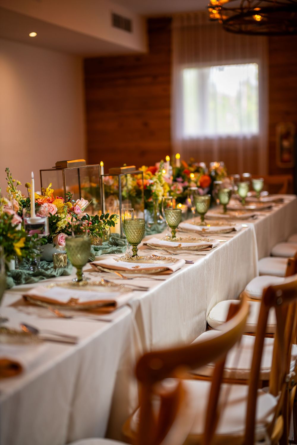 Elegantly set dining table with white linens, wooden chairs and colorful floral centerpieces in rustic reception room.