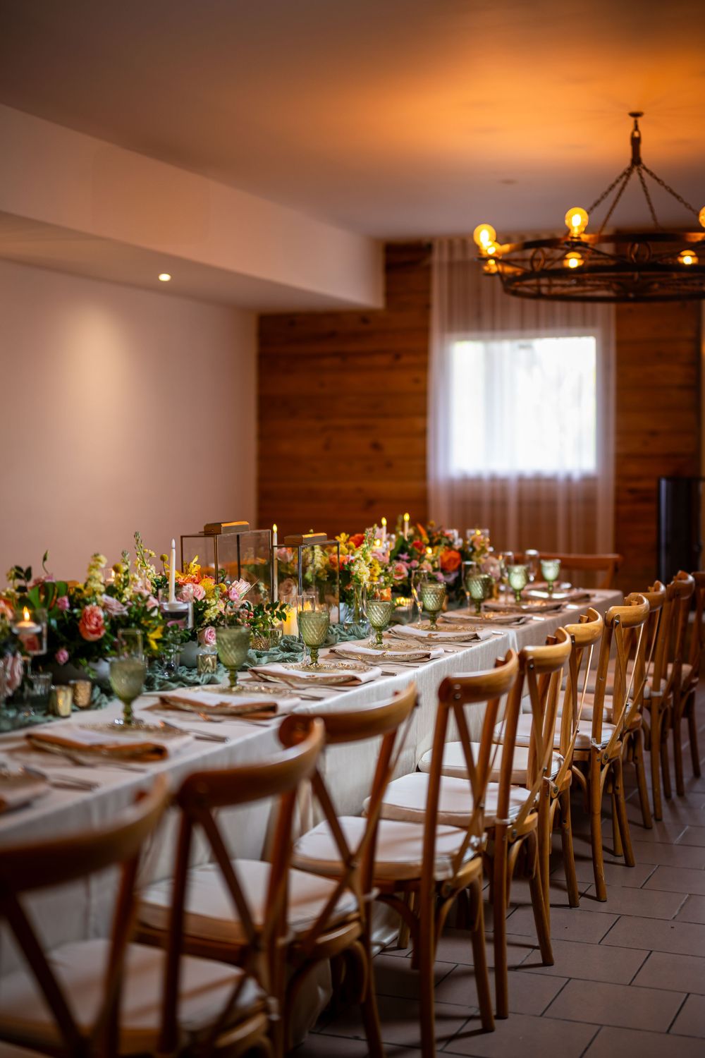 Long banquet table with wooden crossback chairs and floral arrangements under warm chandelier lighting in rustic venue.
