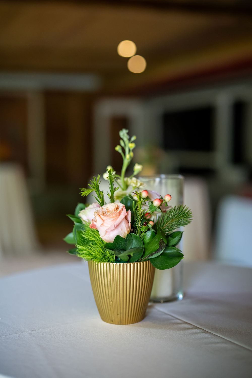 Small gold vase with pink rose and greenery arrangement on white tablecloth.