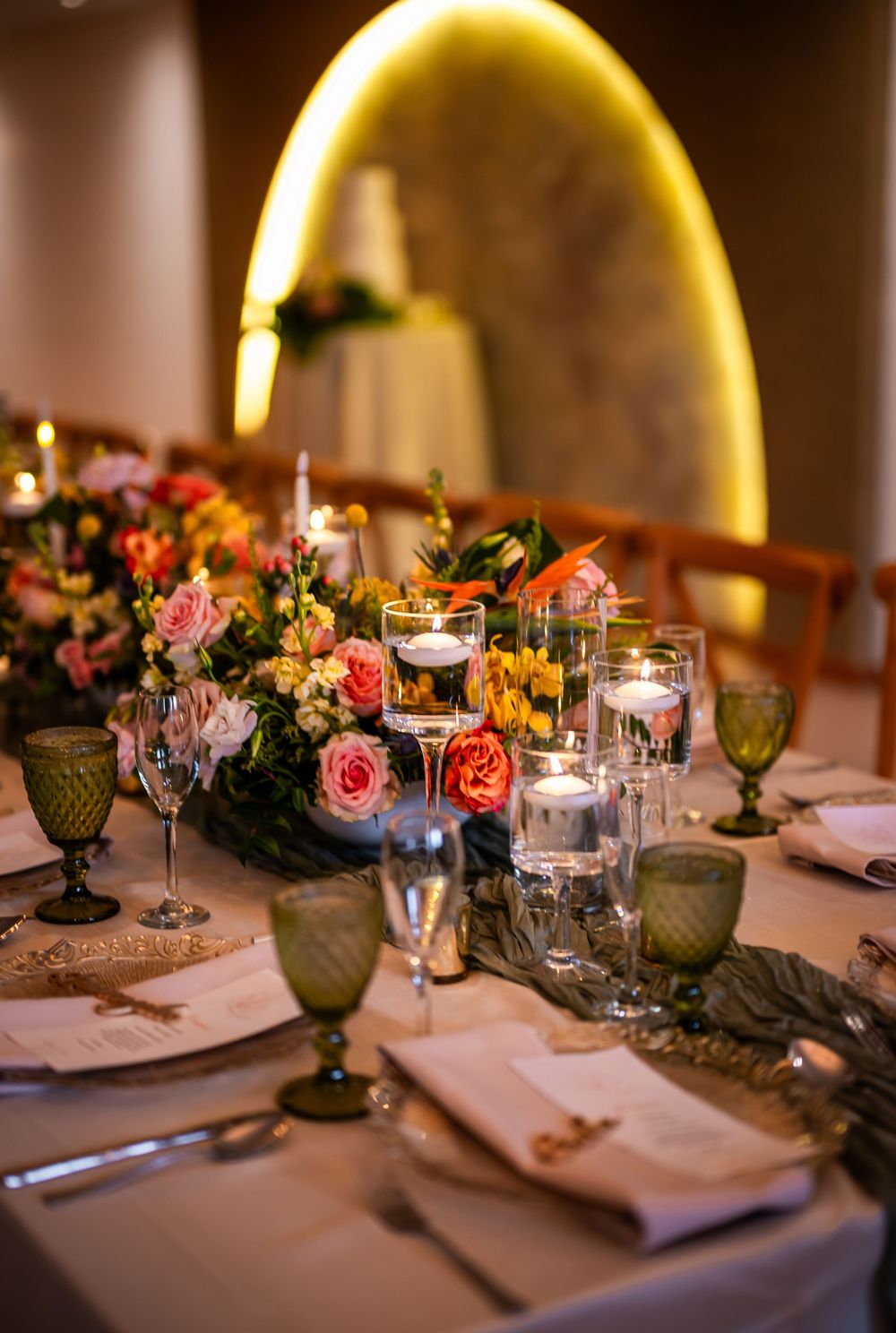 Elegant wedding table setting with pink roses, candles, and sage green goblets under warm ambient lighting.