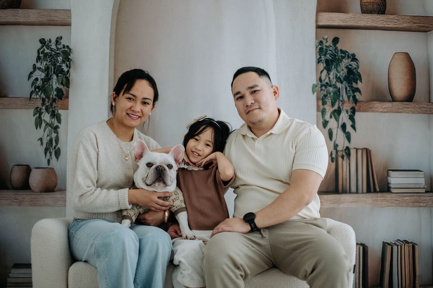 A couple sits on a grey couch with their French Bulldog while indoor plants decorate the modern living room.