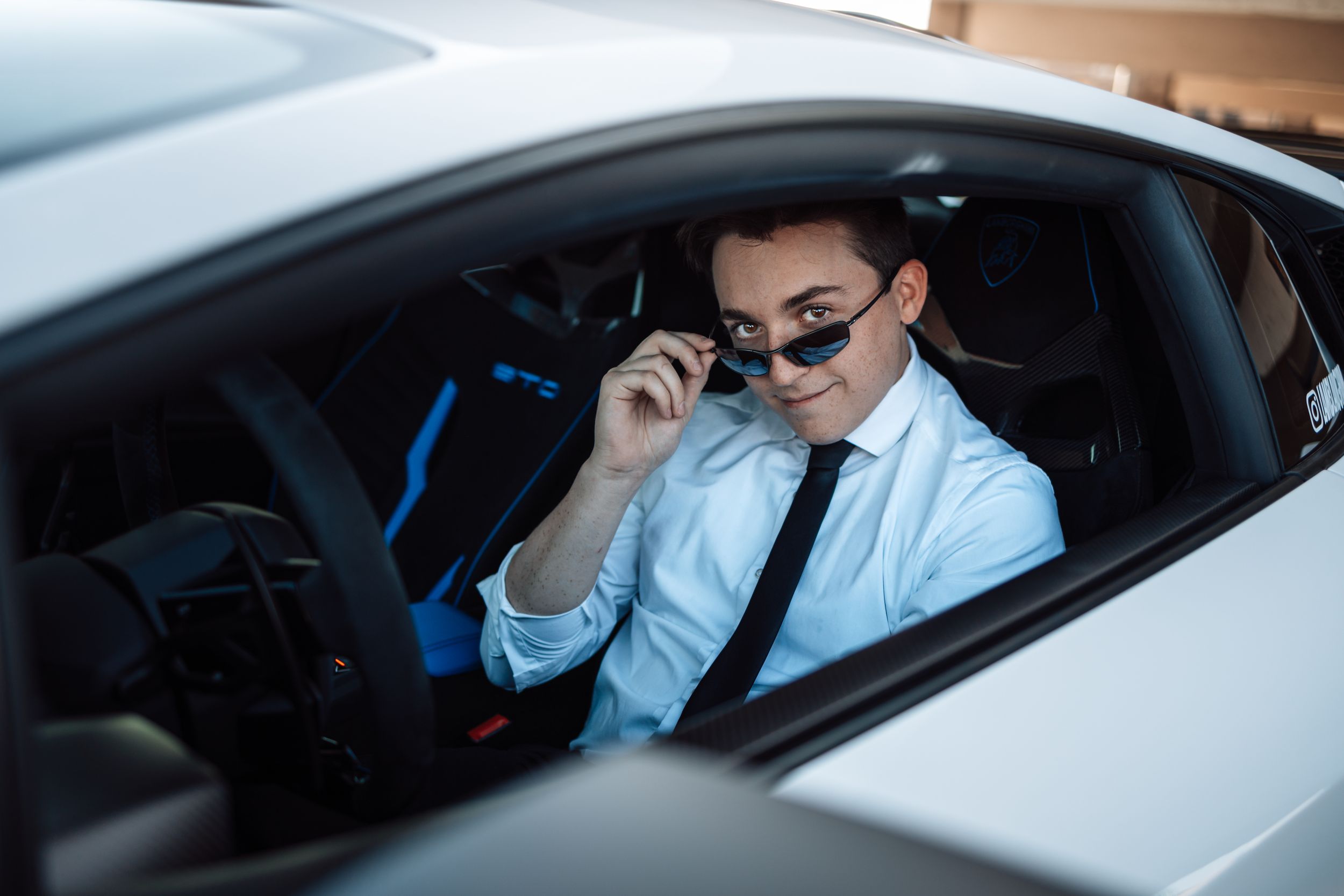 Professional in white dress shirt and black tie adjusts sunglasses while sitting in luxury car.