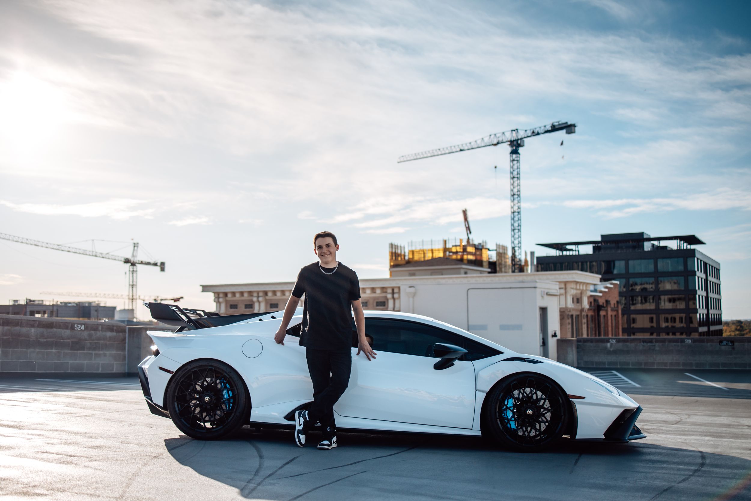White Lamborghini supercar parked on rooftop parking lot with construction cranes and urban buildings in background.