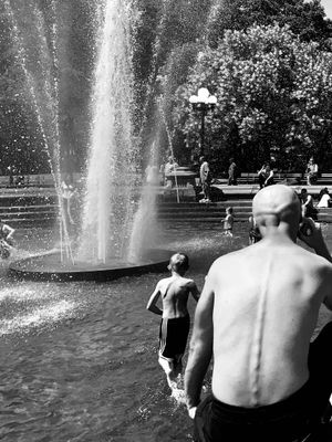 From behind, a father watches his young son run through fountain with water splashing in the back