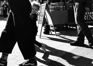 A little kid with a cat face painted on is sneaking behind their mother's leg at a food market