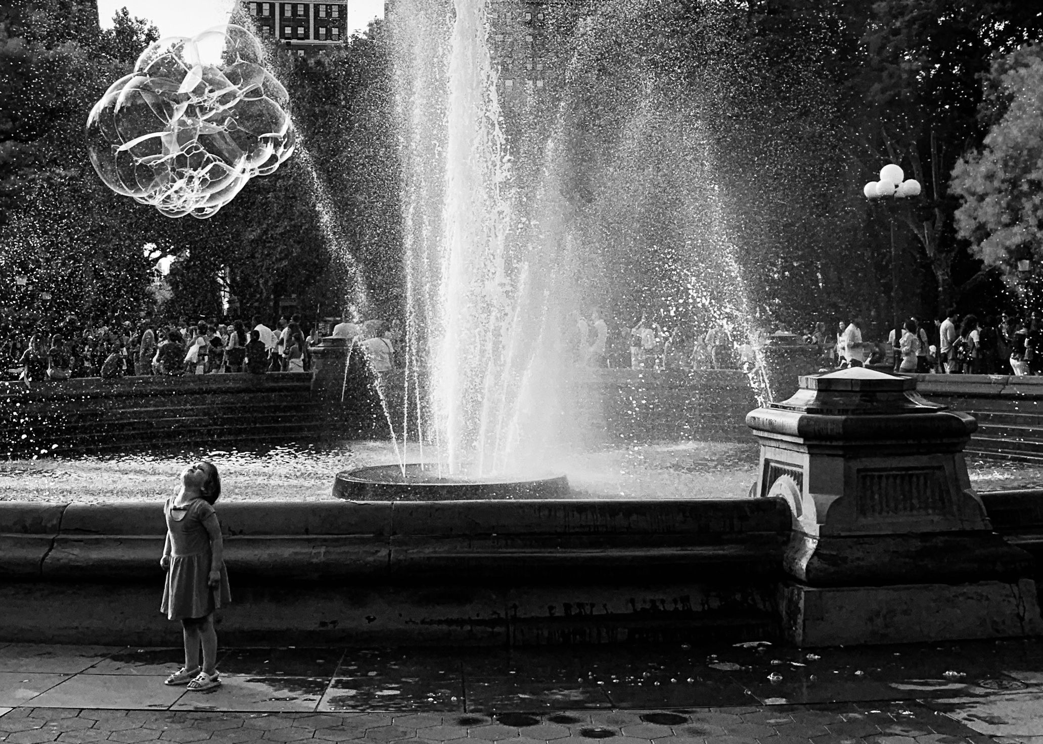 Black and white image of a young girl in a dress standing by the Washington Square Park fountain, watching soap bubbles float in the air