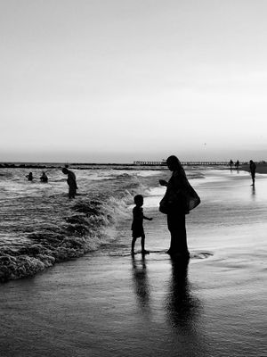 Black and white silhouette of a woman in a hijab standing on the beach with a child