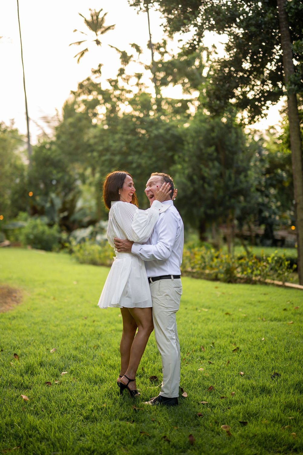 A romantic couple embraces on a lush green lawn at sunset with gardens and palm trees in the background.