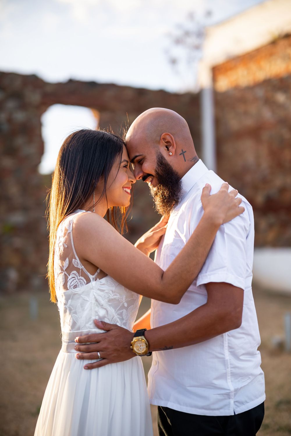 Golden sunlight illuminates a romantic couple in white embracing outdoors against a stone wall background.