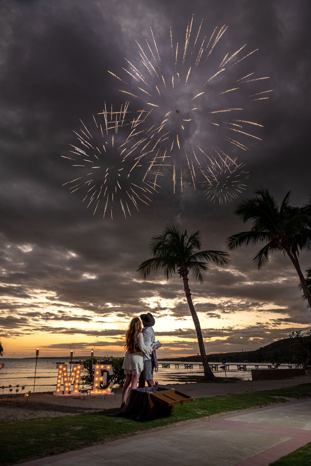 Fireworks burst over a tropical beach at sunset with palm trees silhouetted against a dramatic cloudy sky.
