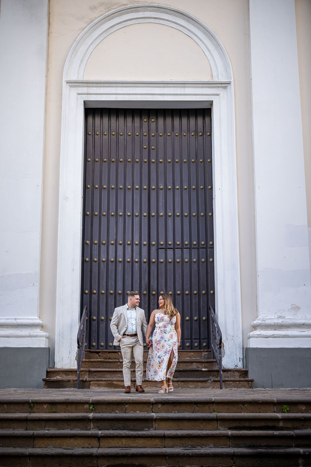 A couple stands on antique stone steps against a white colonial building with large dark wooden doors.