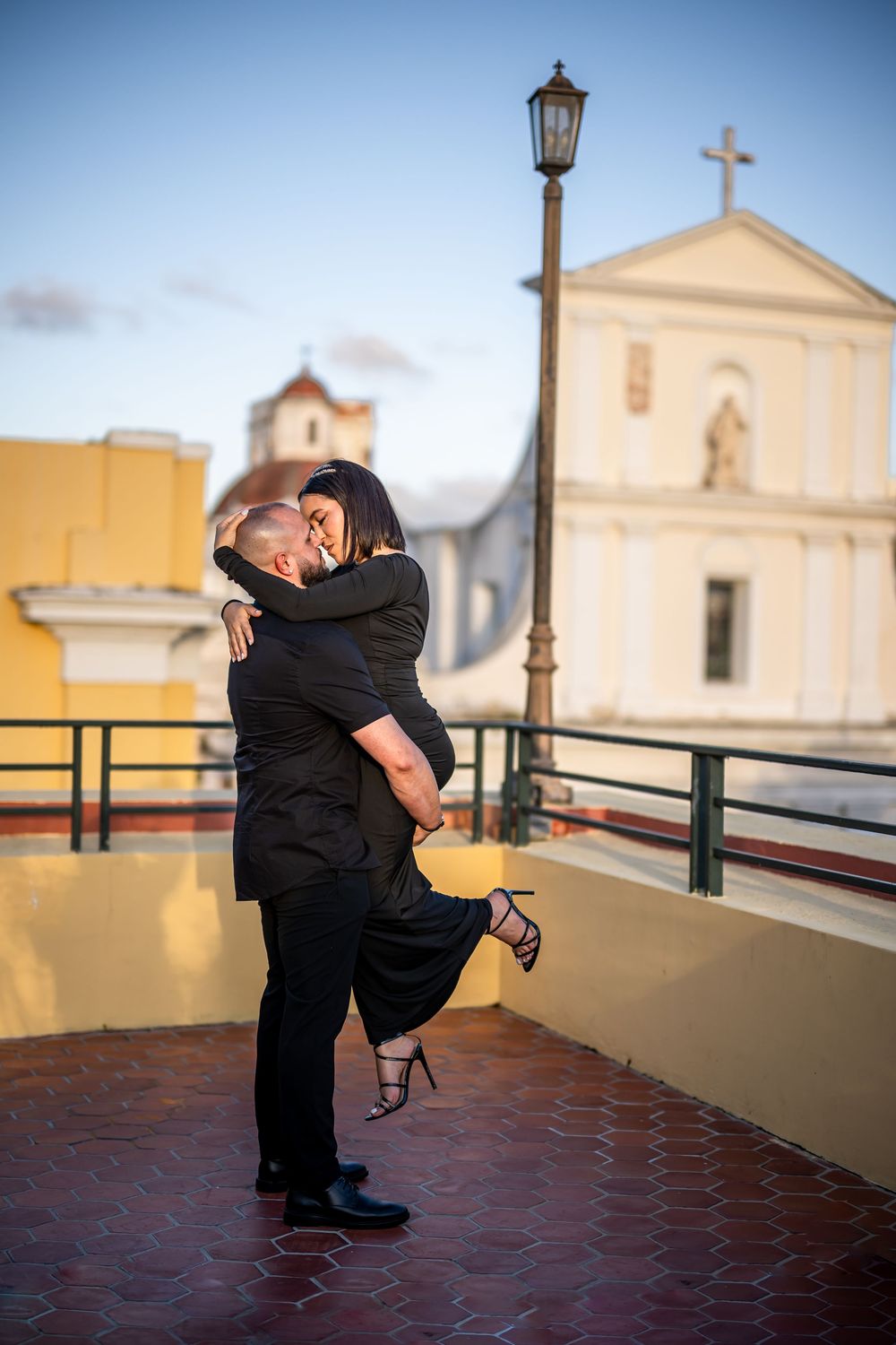 A romantic couple standing together against a church backdrop in dreamy evening light create silhouettes on a tiled terrace.