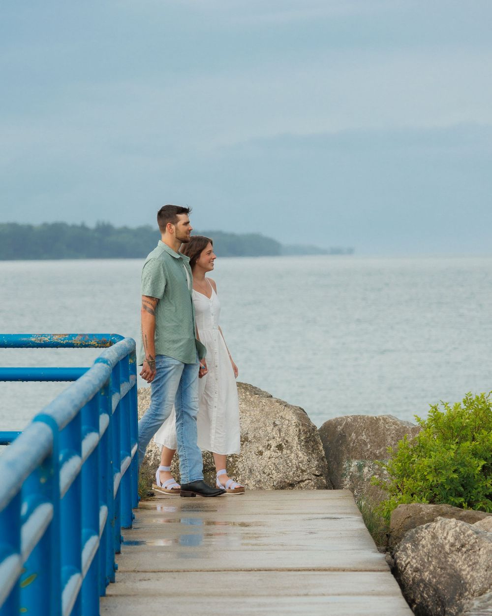 A couple stands together on a seaside walkway with blue railings and rocky shore on a cloudy day.