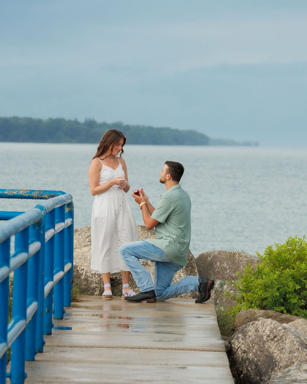 A couple shares a romantic proposal moment on a coastal boardwalk surrounded by rocks and water on a misty day.