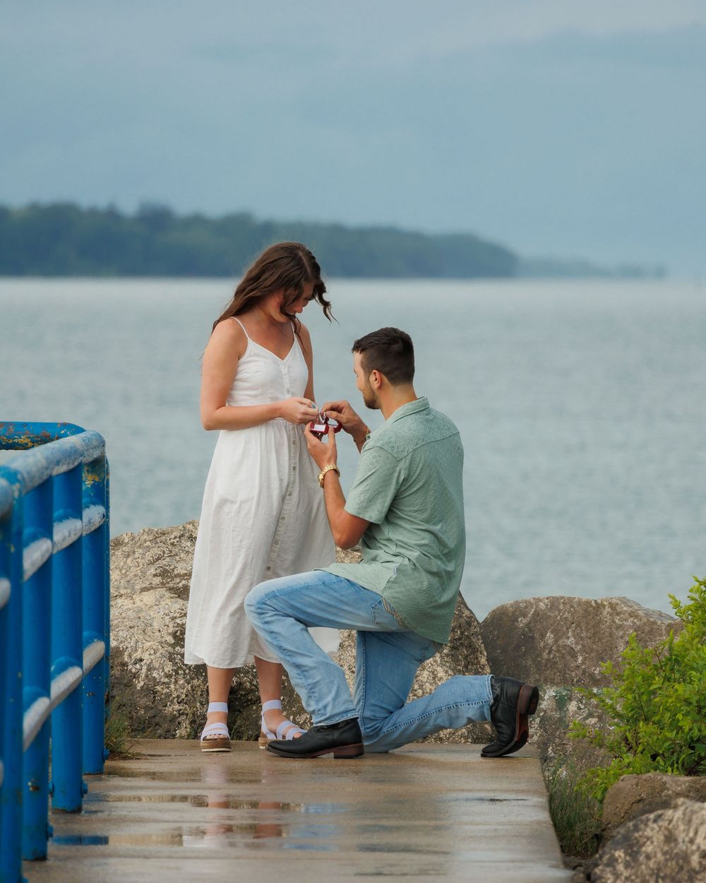 A romantic proposal moment unfolds on a scenic pier overlooking calm waters and distant hills.
