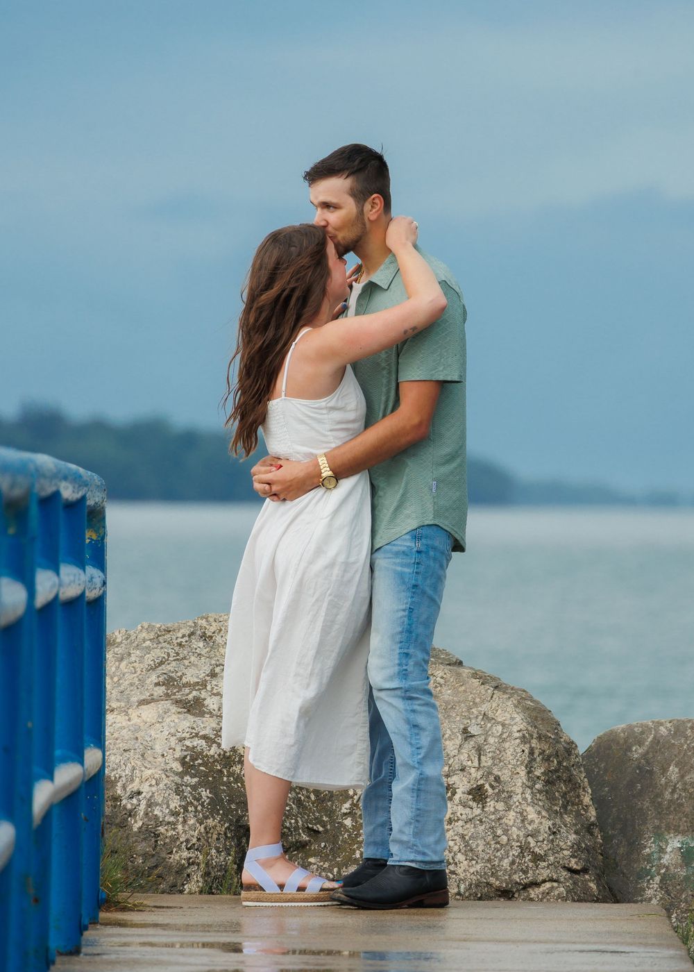 A romantic couple embraces on a coastal walkway with blue railings and ocean view in the background.