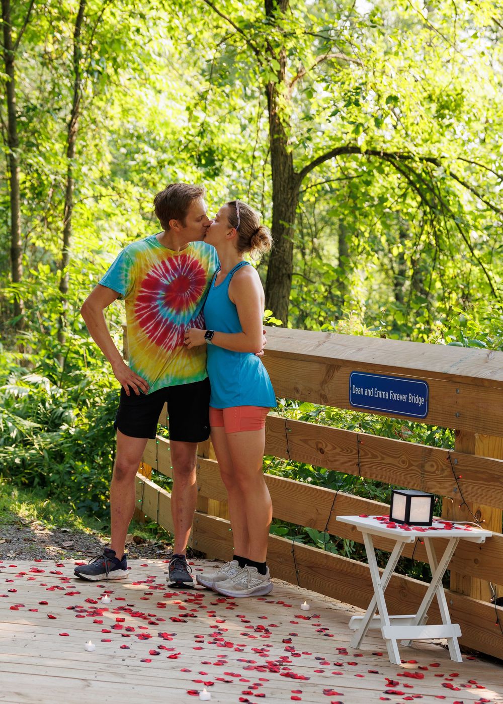 A romantic scene on a wooden deck with red rose petals scattered on the ground and a tie-dye shirt in summer sunlight.