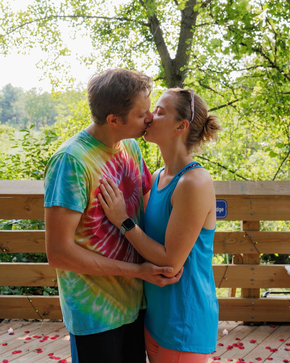 A sweet moment between two people in casual athletic wear sharing a kiss on a wooden deck surrounded by trees.