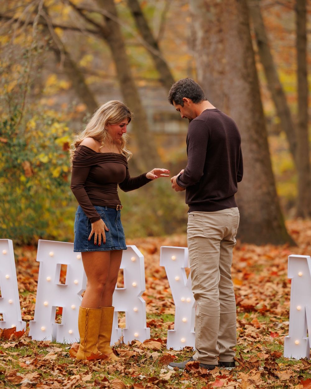 Two people lighting white paper bag luminaries arranged in a row on autumn leaves during a memorial service.