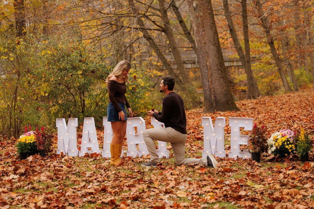 A romantic autumn proposal scene with large white letters spelling MARRY ME surrounded by fall foliage and pumpkins.