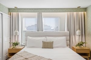 Modern bedroom interior with white bedding, neutral tones, and natural light streaming through horizontal blinds.
