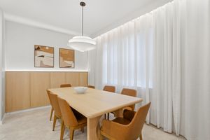 Modern dining room with wooden table, tan chairs, pendant light, and sheer white curtains in a bright space.
