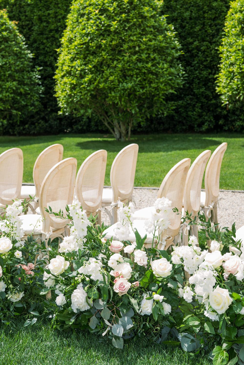 Elegant cream-colored chairs adorned with lush white and pink floral arrangements in a garden setting.