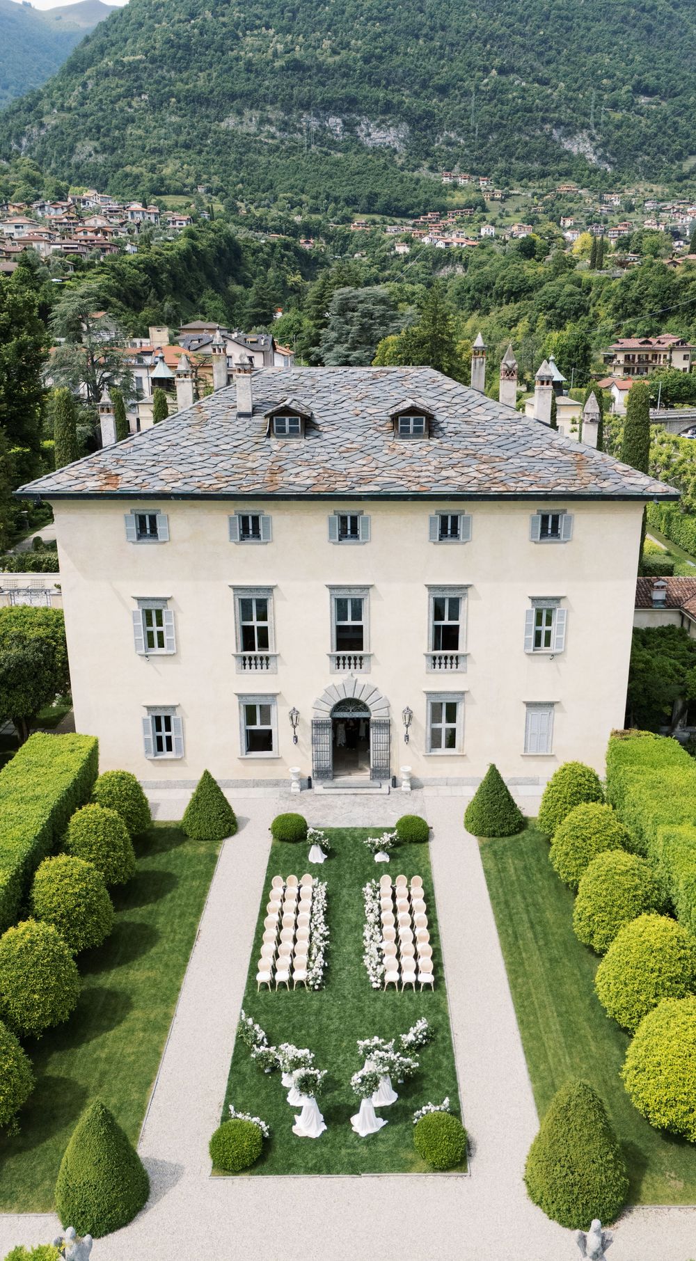 Aerial view of a white villa with symmetrical gardens and ceremony setup surrounded by mountains.