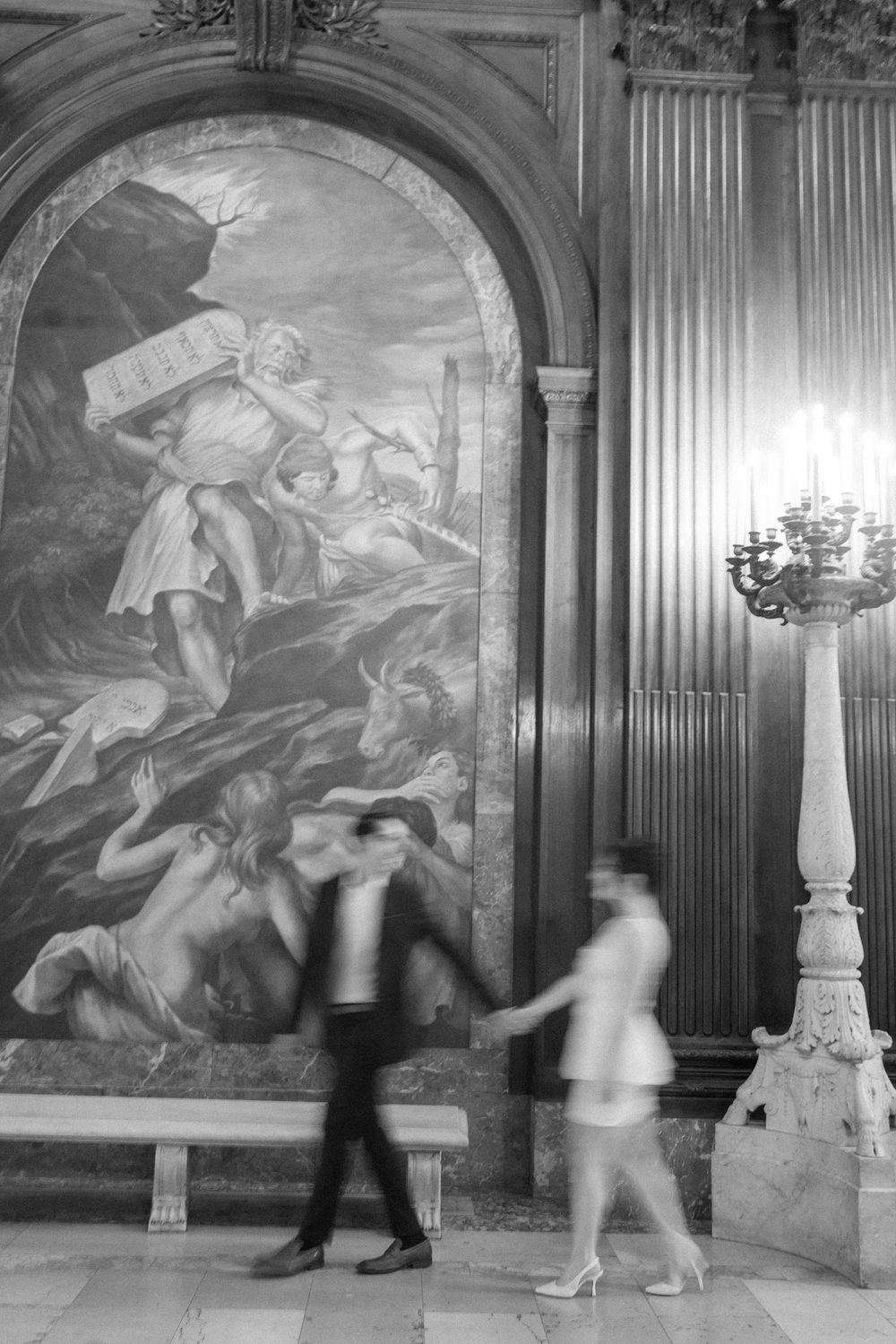 Motion blur of couple walking past dramatic classical mural and ornate candelabra.