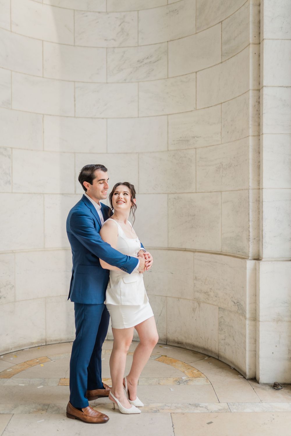 Couple embraces in curved marble alcove wearing formal attire.