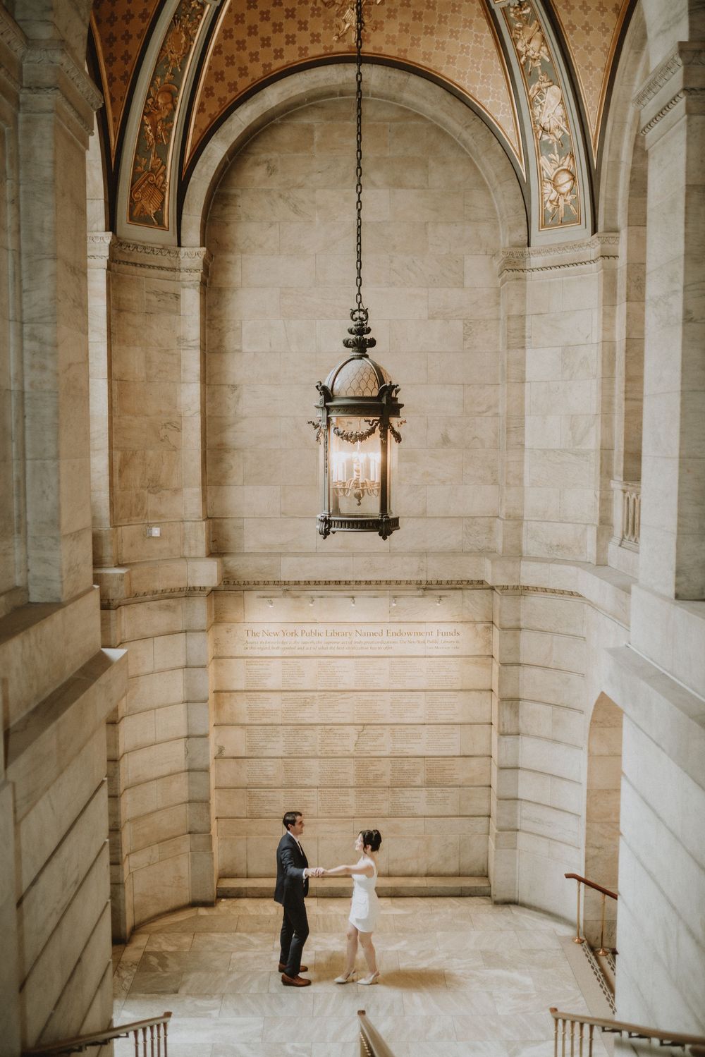 Couple stands beneath ornate hanging lantern in grand marble hallway.