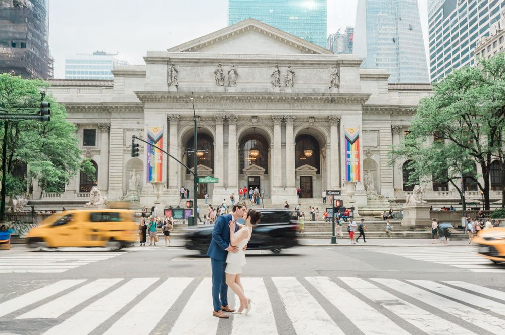 Couple poses in front of New York Public Library as yellow taxi passes by.