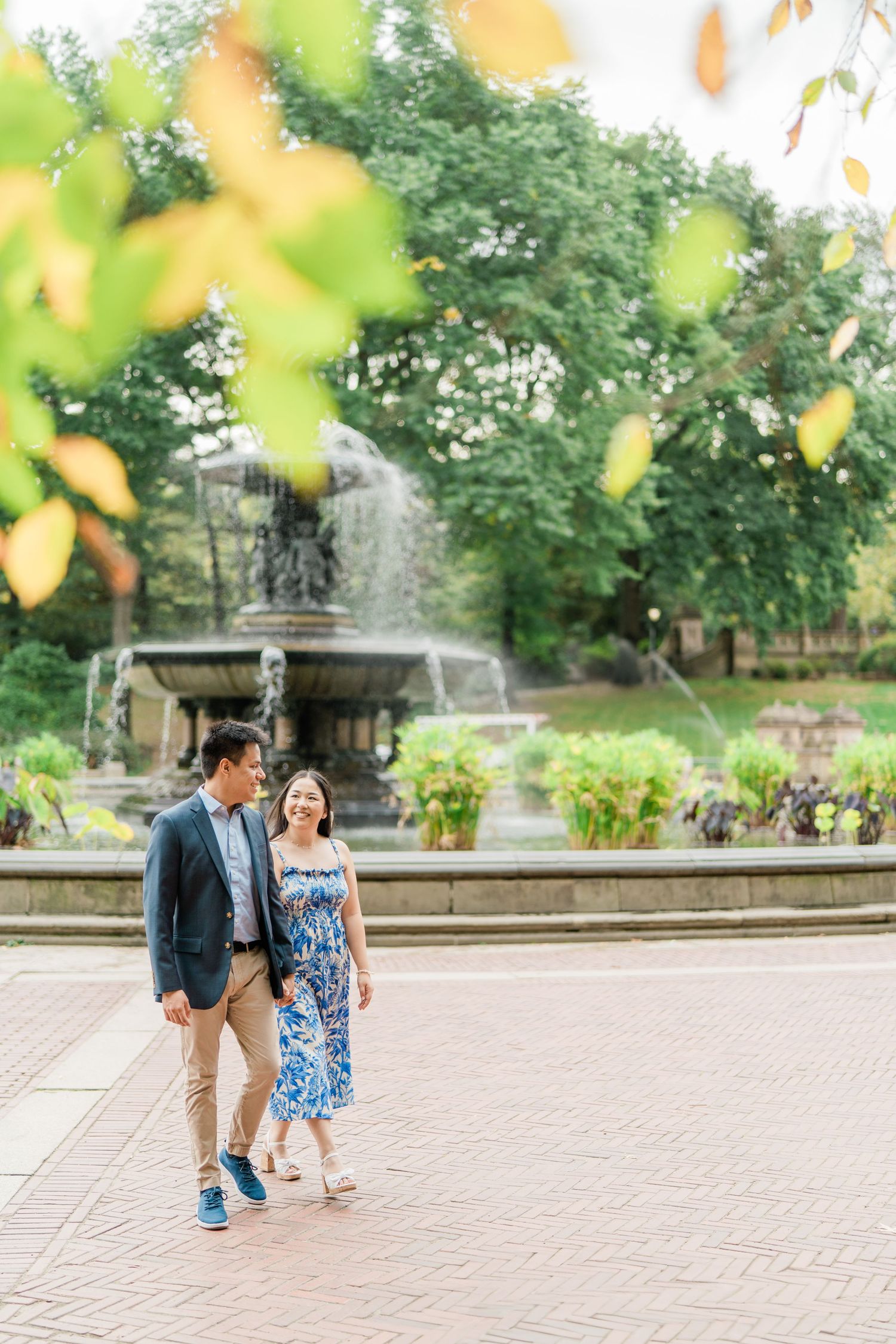 Couple strolling together on a paved pathway surrounded by lush green trees and fountains.