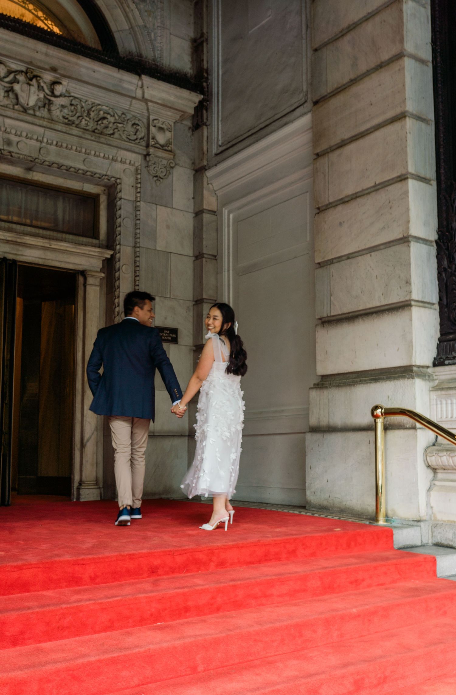 Couple ascending red-carpeted stairs inside an elegant classical building interior.