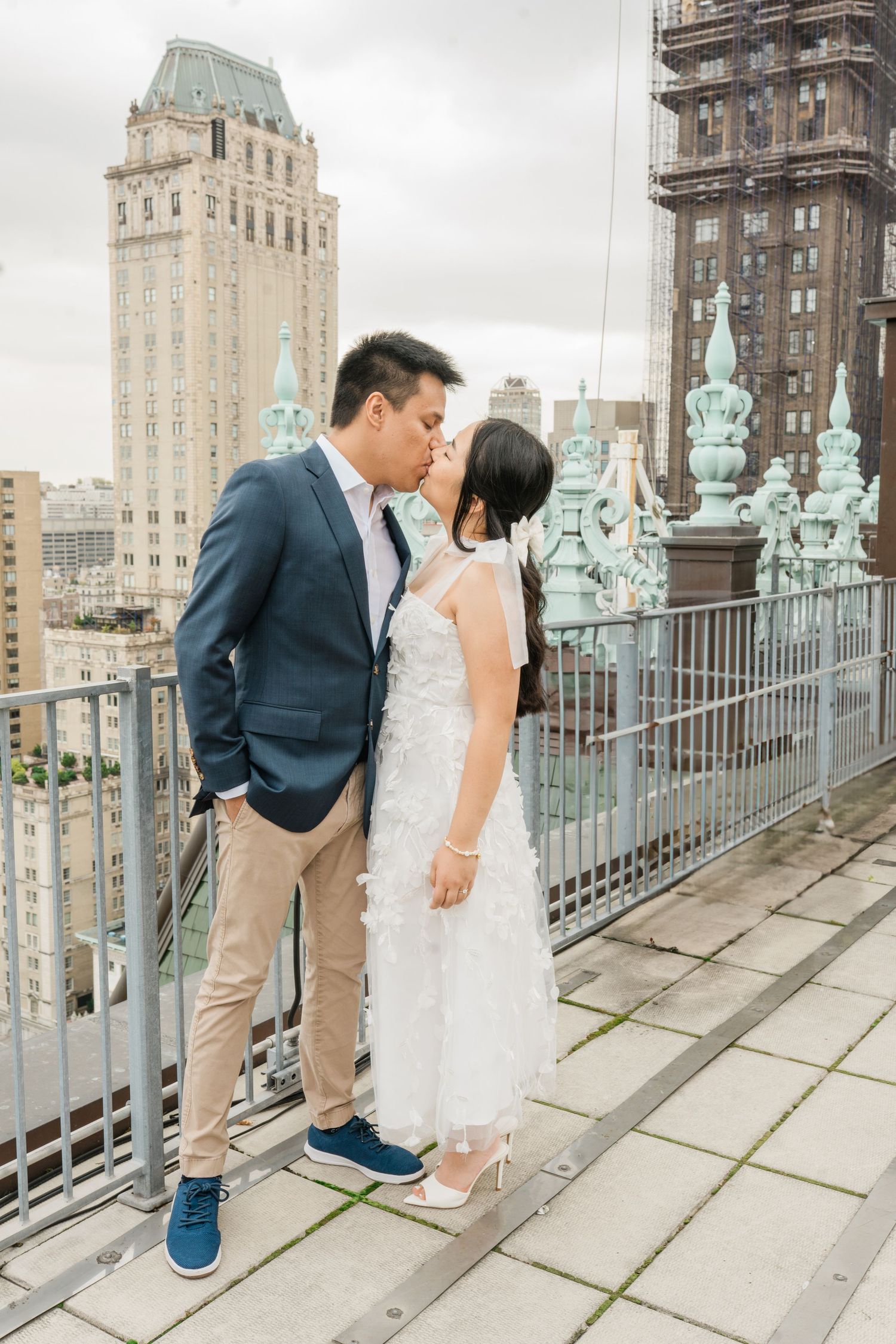 Couple embracing on an ornate balcony with historic New York City architecture in the background.