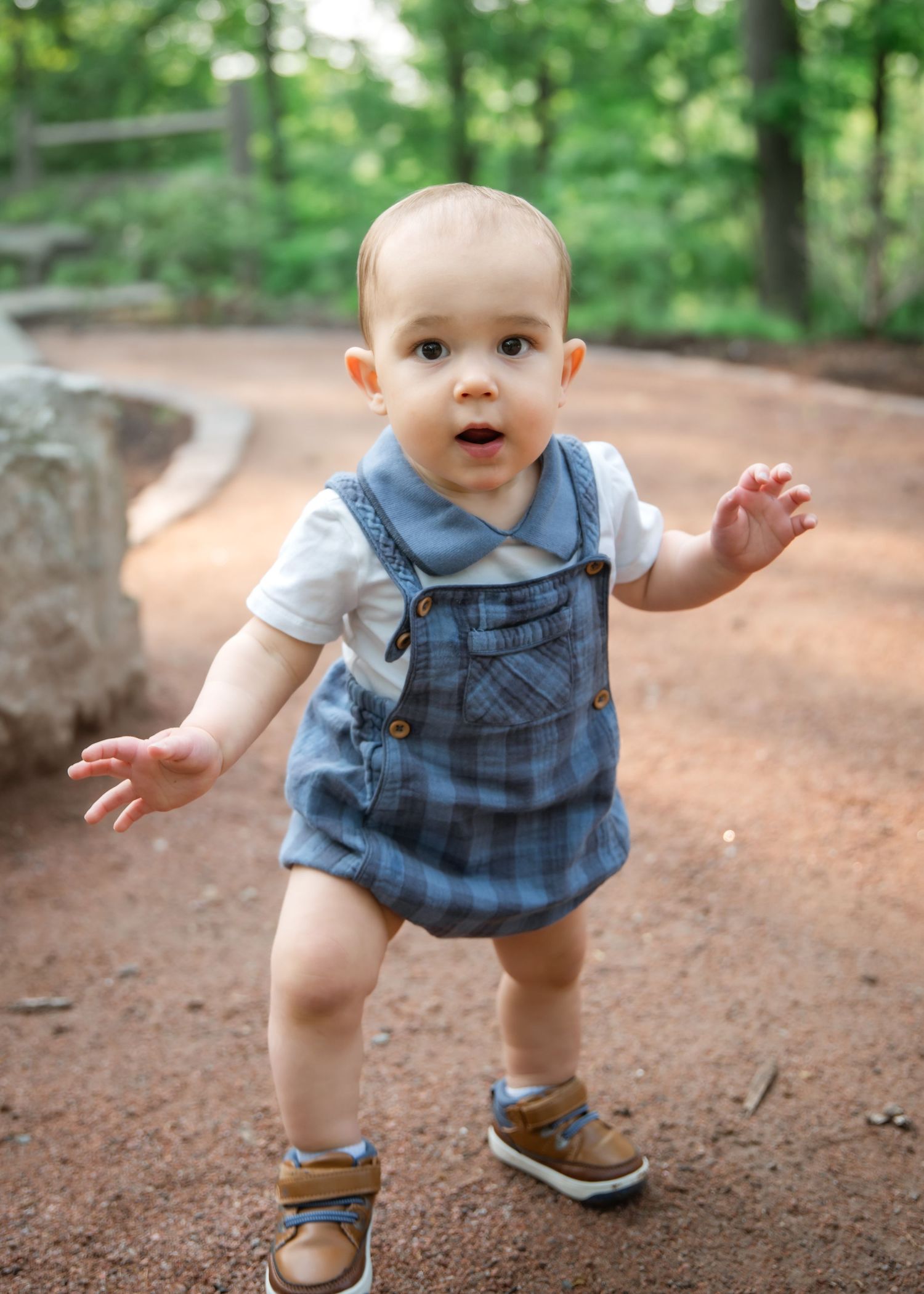 Small child in denim overalls and brown shoes learning to walk on a winding dirt path through a lush green forest.
