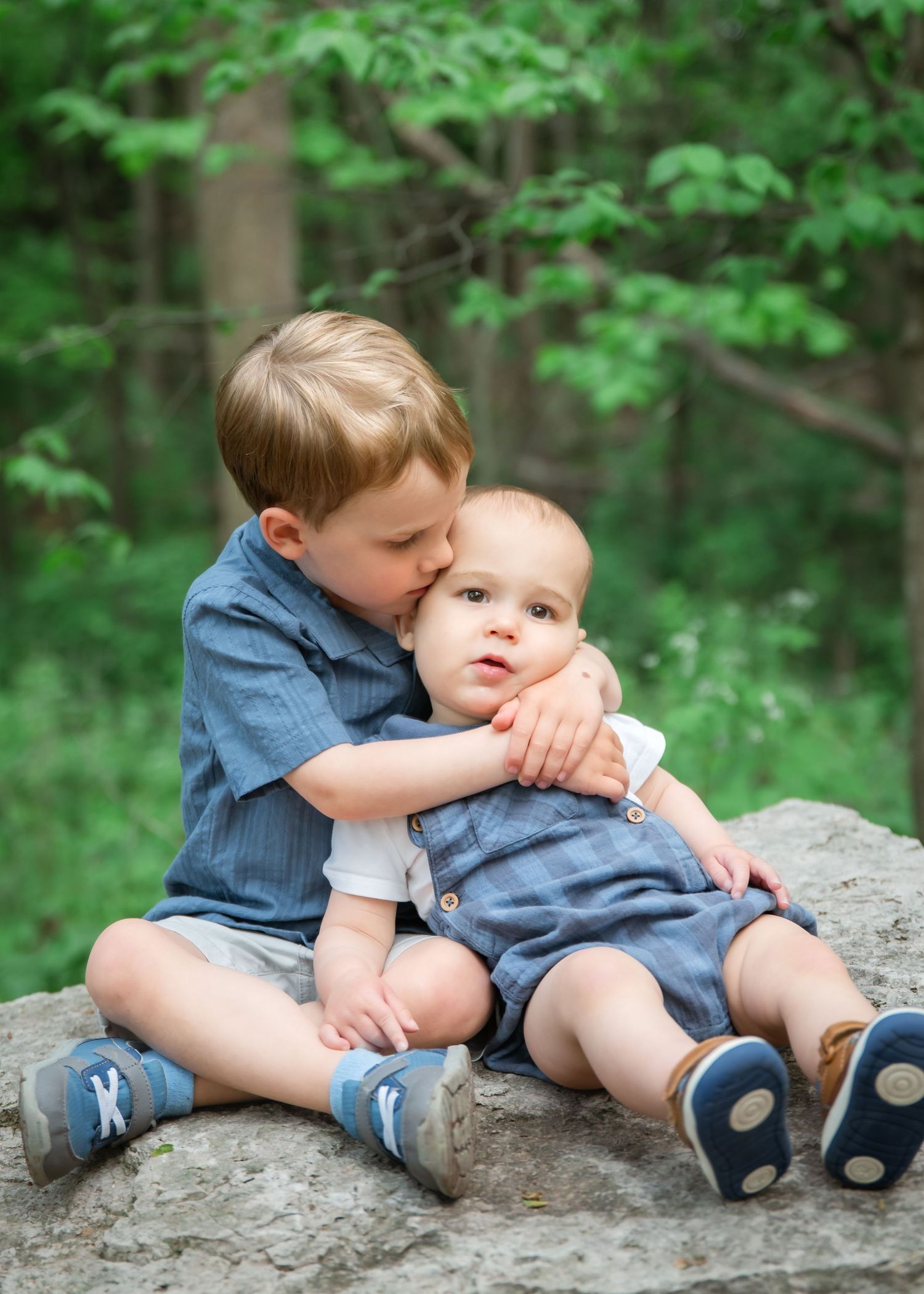 Two young children in denim clothing sit together on a rock in a lush green forest setting.
