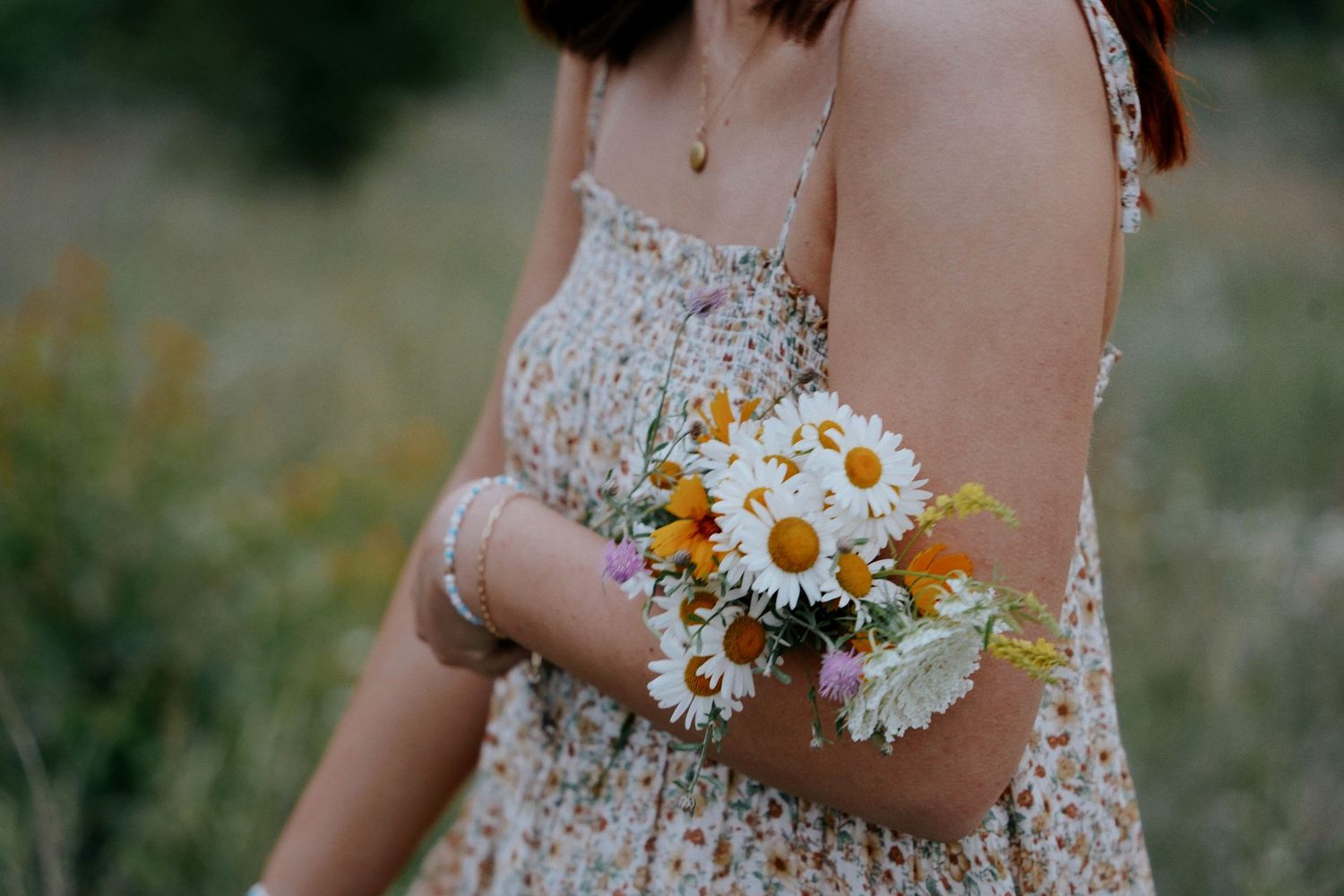 Close-up of woman in floral sundress holding a bouquet of white daisies and wildflowers outdoors.