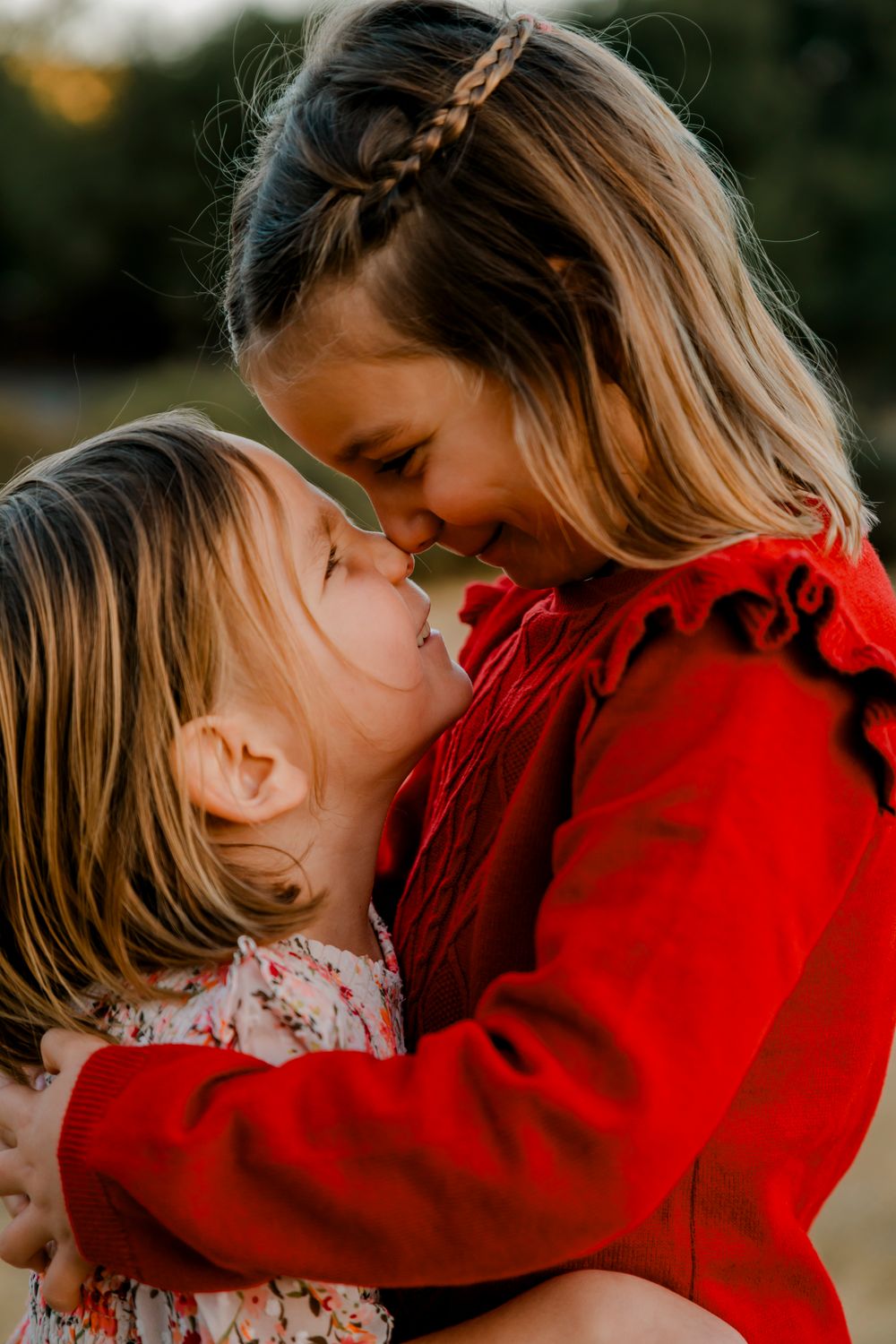 Two girls share a tender moment outdoors wearing red and floral outfits.