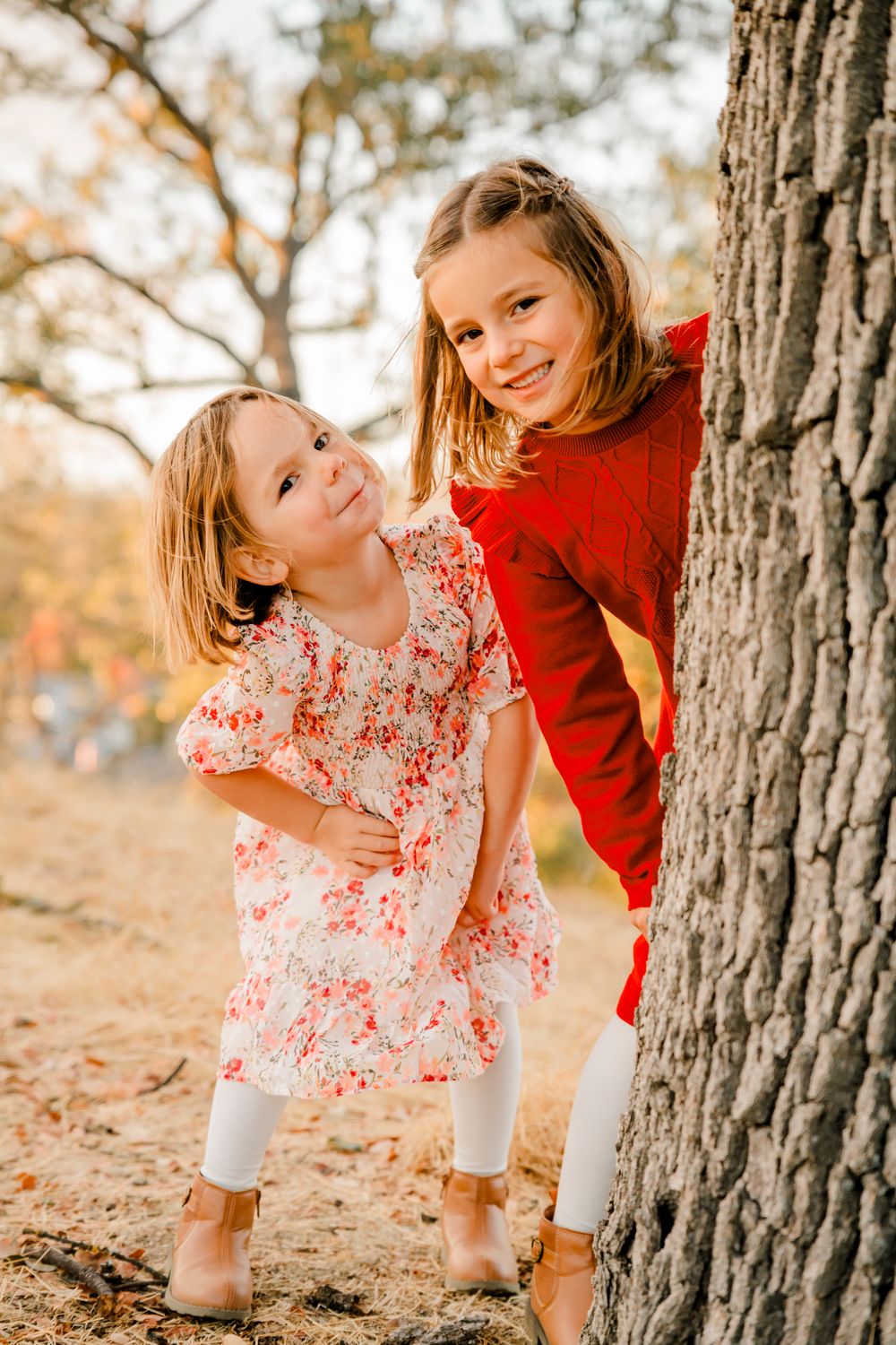 Two children playfully pose by a large tree trunk in autumn clothing.