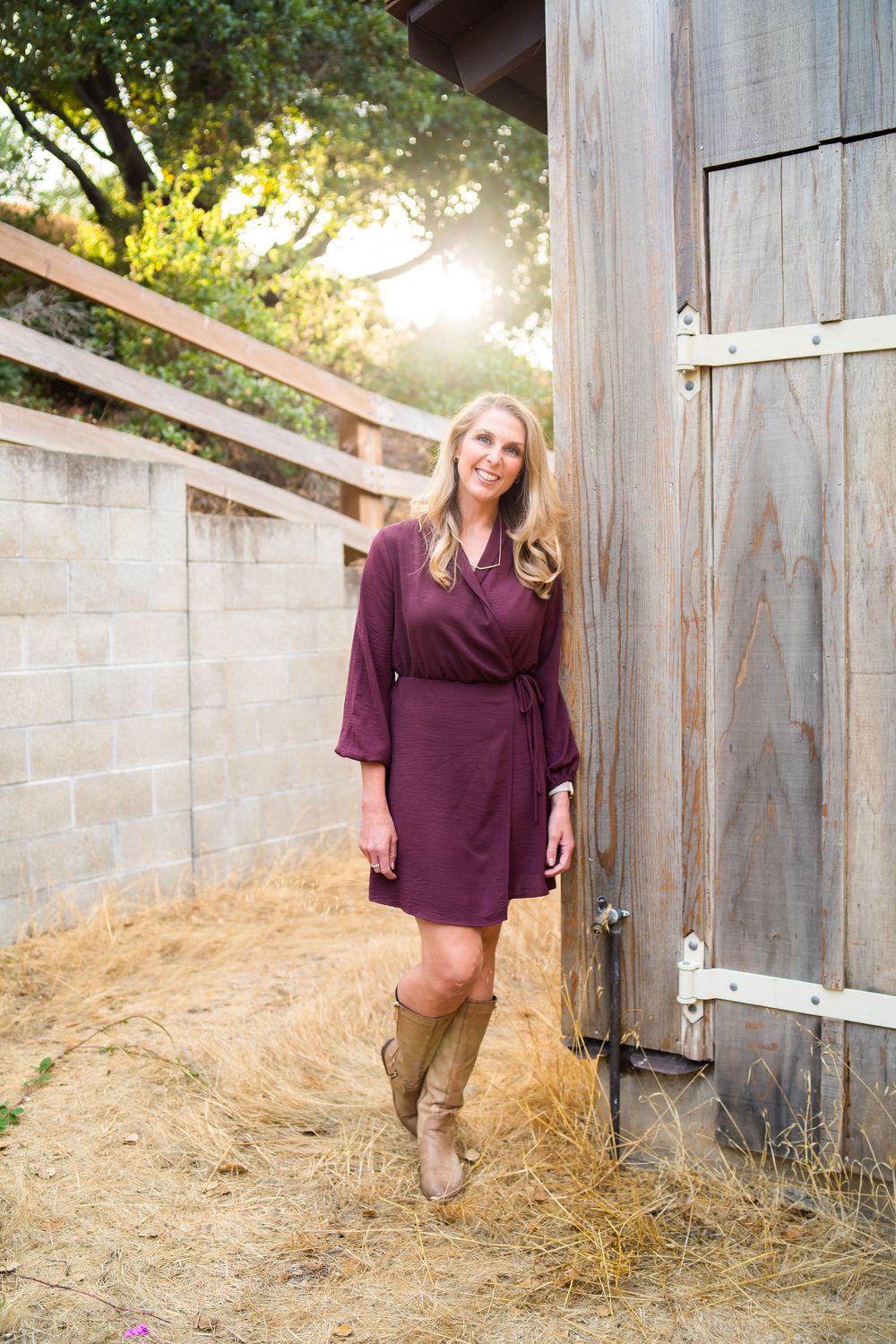 Woman in a burgundy wrap dress and boots posing casually by a rustic barn wall at sunset.