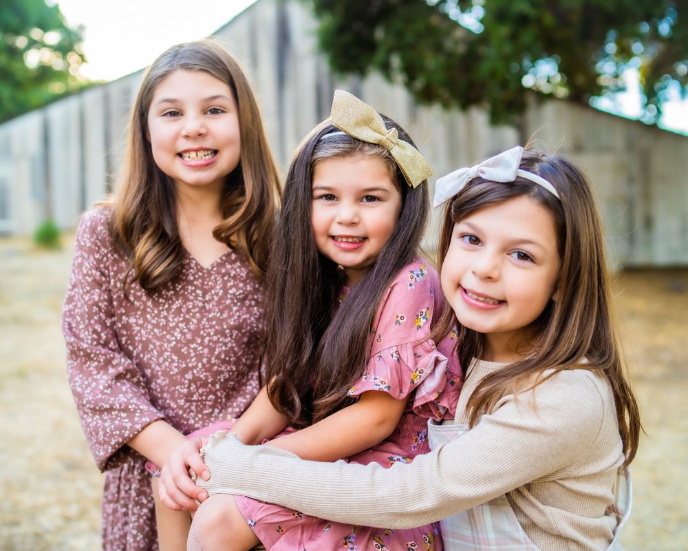 Three young girls in floral dresses pose together outdoors on a sunny day.
