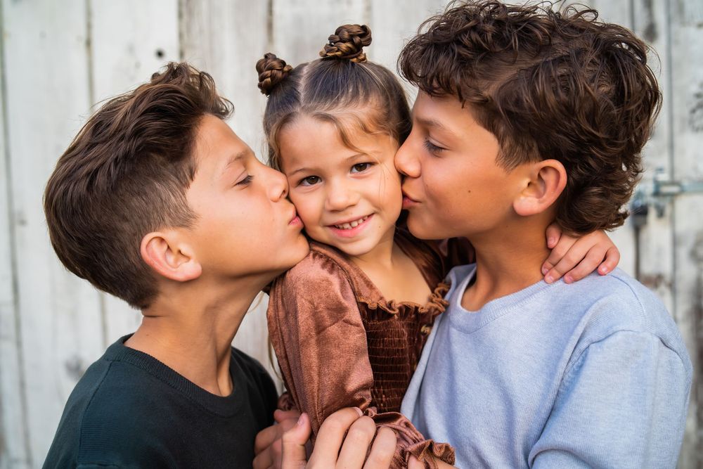 Three children share a sweet family moment together with kisses against a rustic wooden wall background.