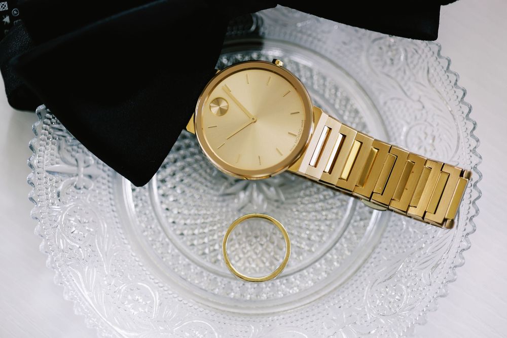 A gold wristwatch and wedding ring displayed on a white lace doily with black fabric in the background.