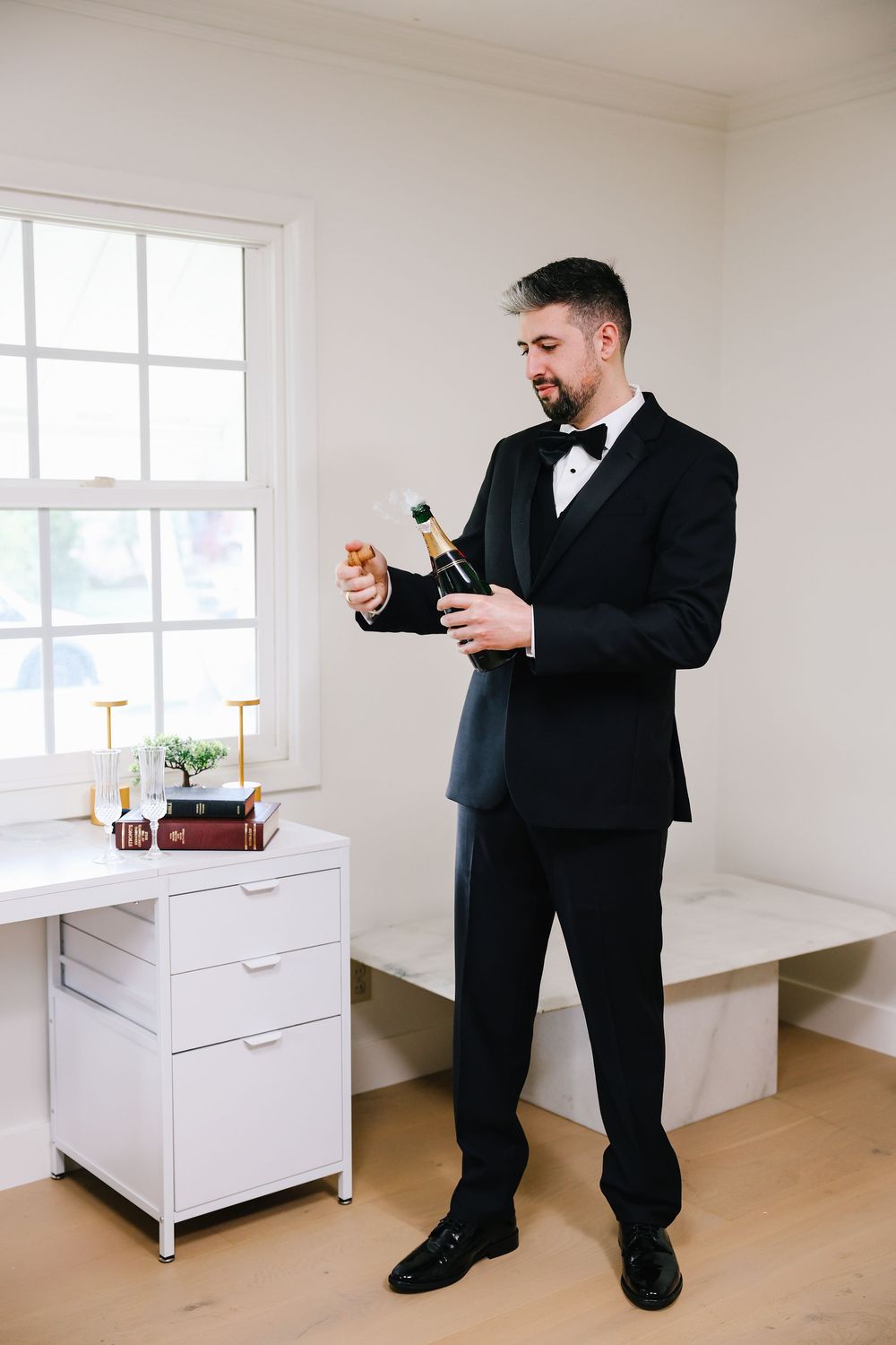 A person in a formal black tuxedo opens a bottle of champagne next to a white cabinet with gold candlesticks.