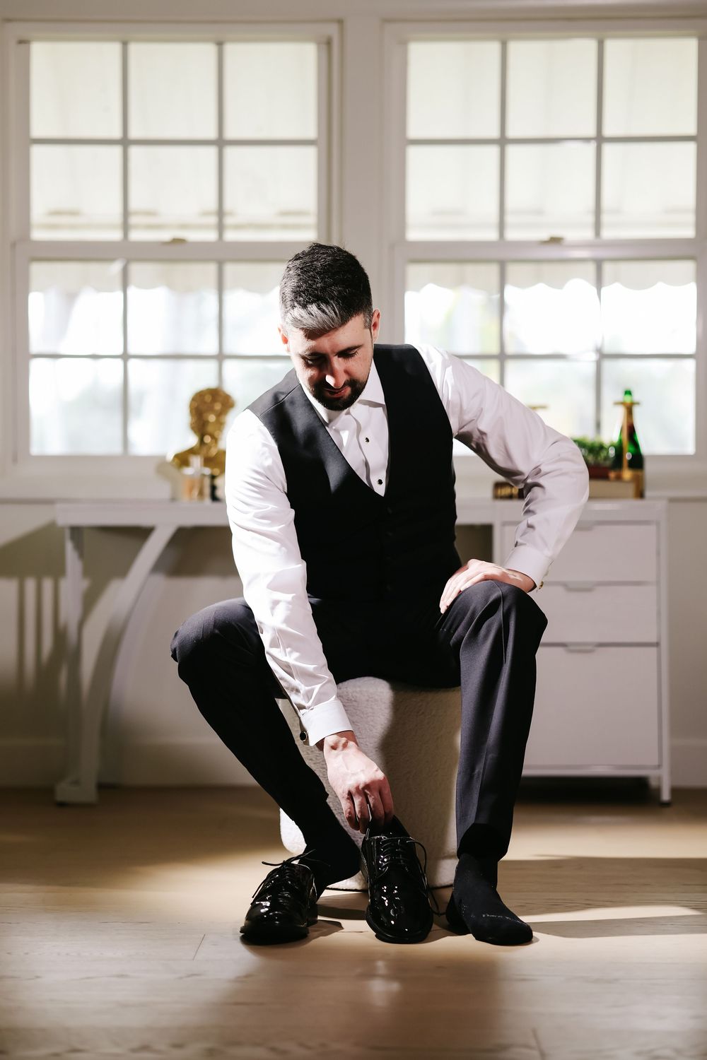 Man in formal vest and white shirt kneels down to tie the laces of dress shoes in a bright room with windows.