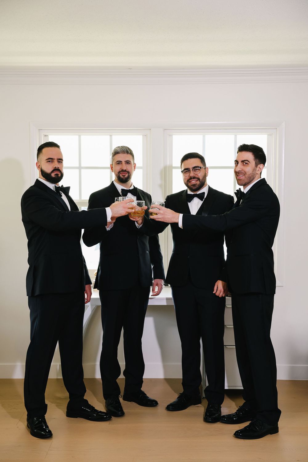 Four groomsmen in black tuxedos and bowties stand together in a bright room with white walls and windows.