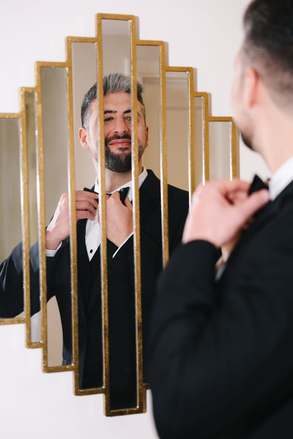 Someone adjusts their bowtie while looking in an art deco style mirror with gold geometric frame design.