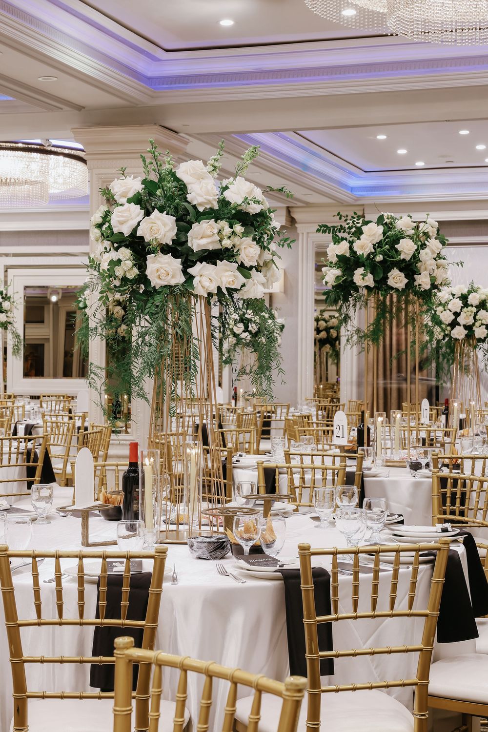 Elegant ballroom wedding reception setup with tall white floral centerpieces, gold chiavari chairs and crystal chandeliers.